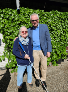 Eine ältere Frau und ein älterer Herr stehen nebeneinander in einem Garten mit bewachsener Hecke im HIntergrund. Die Frau hält einen Blindenlangstock.