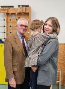 ein älterer Mann und eine junge Frau stehen in einer Turnhalle. Die Frau hält ein Kind im Arm, das den Rücken zur Kamera gedreht hat. Die Mutter lächelt in die Kamera.