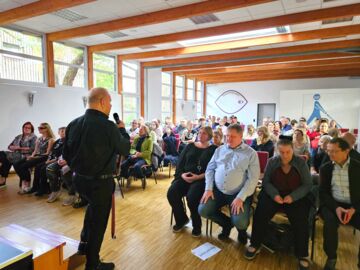 Ein mit Menschen gut gefüllter Saal ist zu sehen, sie alle sitzen aufmerksam. Im Vordergrund steht ein Mann in der Hinteransicht, er hält ein Mikrofon in der Hand.