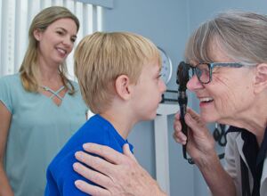 Eine Augenärztin untersucht einen Jungen im Grundschulalter mit einem Ophthalmoskopiegerät (Betrachtung des Augenhintergrunds, Retina und Pupille)