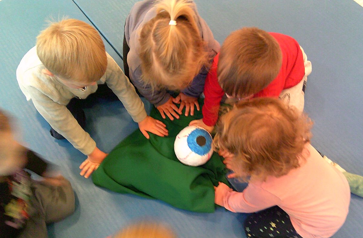 Vier kleine Kinder beugen sich über ein gebasteltes großes Auge, das in einer grünen Decke eingewickelt ist.